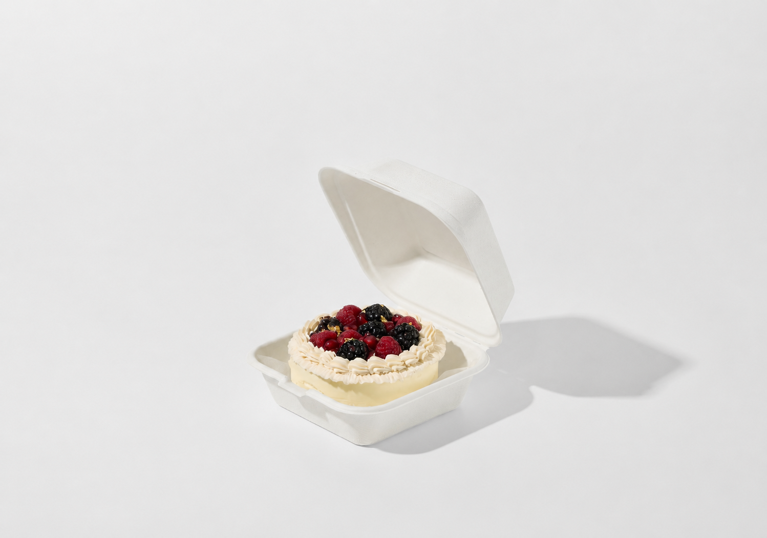 A small open white sugarcane take-out container holding a cream-colored dessert topped with fresh berries, photographed on a clean white background with a soft shadow.