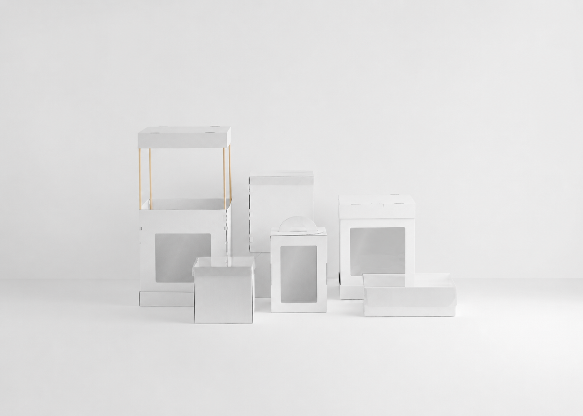 A set of minimalist white cardboard cake boxes packaging boxes and display stands with clear window cutouts, arranged on a seamless white studio background.