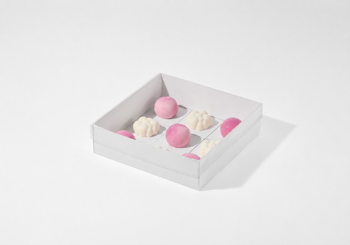 A white square gift box containing several pink and off-white chocolates, photographed on a clean white background with a soft shadow.